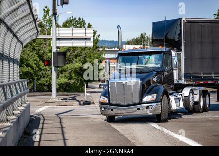 Black stylish day cab big rig semi truck tractor for local deliveries transporting cargo in covered dry van semi trailer turning with front wall spoil Stock Photo