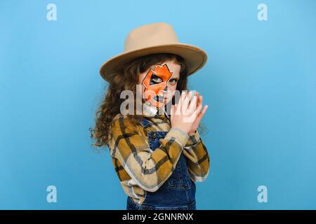 Portrait of little girl child with makeup mask wears brown hat says Boo laughs, spooky smiling looking at camera, posing isolated over blue color wall Stock Photo