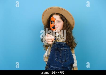 Portrait of girl kid with Halloween makeup mask wears hat and shirt, point index finger camera on you motivation encourage, posing over blue wall Stock Photo