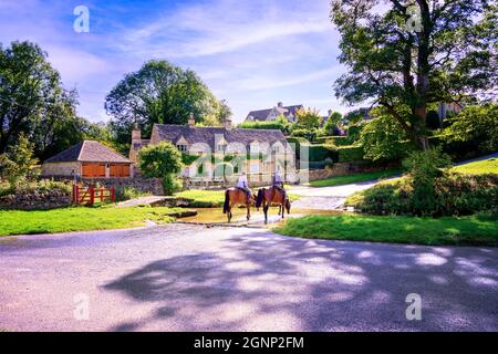 Two horses & riders crossing ford across the river traditional stone built Cotswold village cottage in background. Upper Slaughter Gloucestershire UK Stock Photo