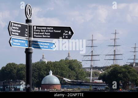 A riverside signpost points along the Thames Path between Tower Bridge, the Thames Barrier and the Cutty Sark cargo clipper whose tall masts are seen near the twin domes of Greenwich Hospital, on 16th September 2021, in London, England. Stock Photo