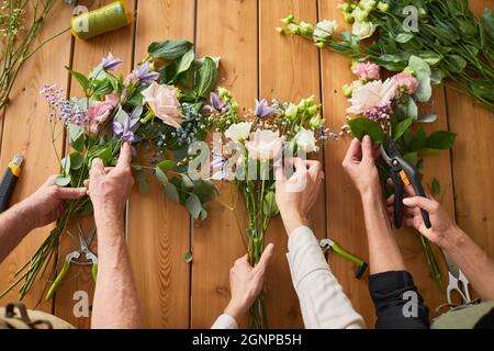 Top view closeup of three florists arraigning flower compositions on wooden table in cozy workshop, copy space Stock Photo