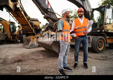 Two men engineers discussing their work standing against construction machines Stock Photo