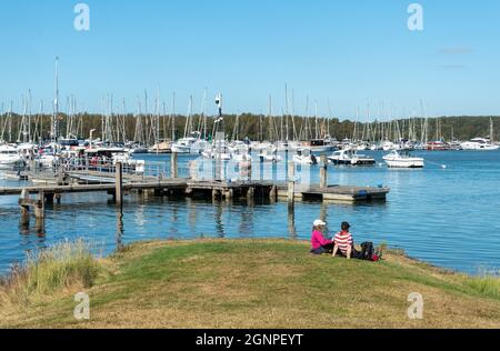 People sitting and enjoying a sunny day watching the boats and yachts on Beaulieu River from Bucklers Hard village in the New Forest, Hampshire, UK Stock Photo