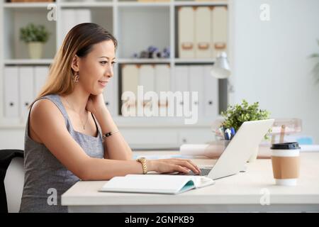 Content young Vietnamese businesswoman sitting at table in office and analyzing information on laptop Stock Photo