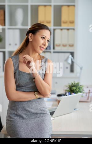 Pensive Vietnamese female executive in gray dress leaning on table with office supplies and looking away Stock Photo