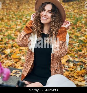 Portrait of beautiful young woman with curly hair posing on the street with autumn leaves on blurred background. Joyful woman looking at camera and smiling while touching elegant hat. Stock Photo