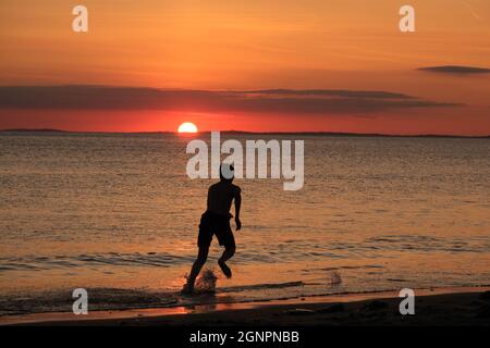 Gower, Swansea, UK. 24th August 2021.  UK weather:  A boy enjoys skim boarding the shallows under a setting sun on a dry, fine  and sunny evening at Llangennith beach on the Gower peninsula. The outlook for the next few days is for similarly fine weather with some warm spells.Credit: Gareth Llewelyn/Alamy Stock Photo