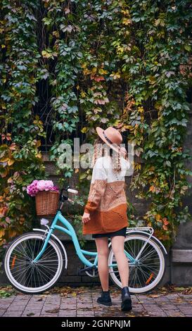 Rear view of curly lady in hat and sweater nearby her blue women's street bike parked against gray wall of house, which almost full covered with vegetation green. House overgrown by green ivy. Stock Photo