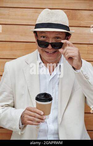 Portrait of happy mature Asian man in white jacket and stra hat taking off sunglasses and drinking coffee outdoors Stock Photo