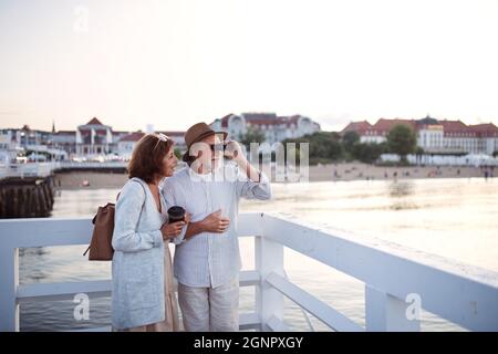 Happy senior couple hugging outdoors on pier by sea, looking at view through binoculars. Stock Photo