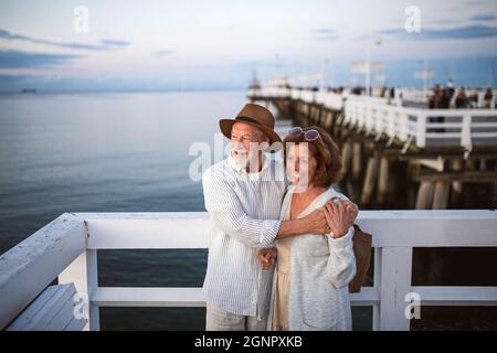 High angle view of happy senior couple hugging outdoors on pier by sea, looking at view. Stock Photo