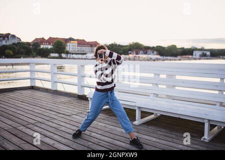 Portrait of preteen girl taking photo with camera on pier by sea at sunset, holiday concept. Stock Photo