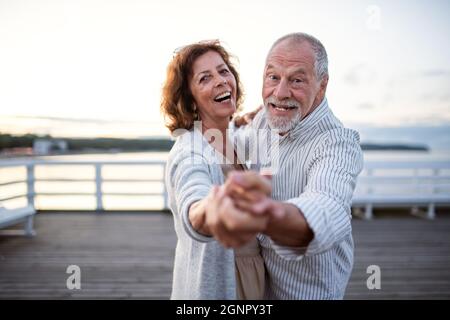 Happy senior couple dancing outdoors on pier by sea, looking at camera. Stock Photo