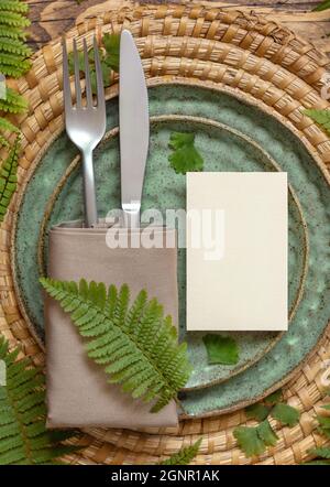 Blank paper card on table setting decorated with fern leaves on wooden table top view. Tropical mock-up scene with place card flat lay Stock Photo
