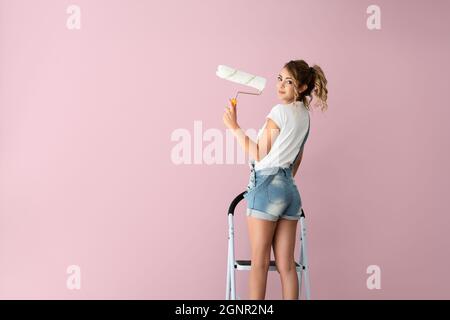 Beautiful woman is standing on the ladder and is painting the wall to pink color with a roller on her hand. High quality photo Stock Photo