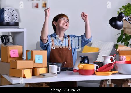 Successful mature Asian woman entrepreneur, Business owner with arms up while wooking with laptop computer at home office with her clay ceramic produc Stock Photo