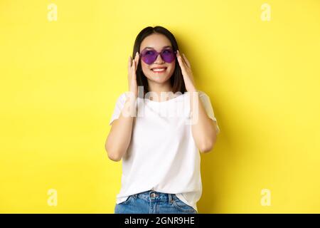 Fashion and lifestyle concept. Stylish asian woman trying new sunglasses in heart-shape, going on vacation, smiling happy at camera, standing over Stock Photo