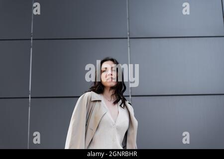 young stylish woman in raincoat looking at camera near grey wall Stock Photo