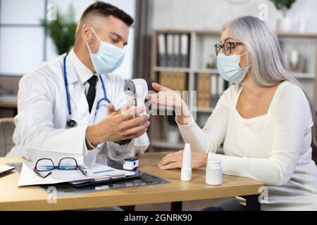 Male physician in face mask and lab coat using electronic contact less thermometer while examining health condition of retired woman. Health care during pandemic. Preventive measures. Stock Photo