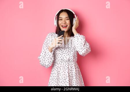 Beautiful smiling asian woman singing song in smartphone microphone, playing karaoke app and using headphones, standing over pink background Stock Photo