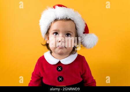 Smiling Portraite Cute Cheerful Chubby Baby Girl in Santa Hat Looking Up Interested At Yellow Background. Child Play Christmas Scene Celebrating Stock Photo