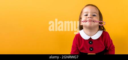 Banner Portraite Cute Cheerful Chubby Baby Girl in Santa Suite Holding Eating Caramel Candy At Yellow Background. Child Play Christmas Celebrating Stock Photo