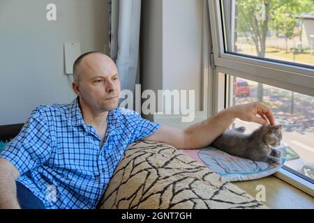 portrait of a mature pensive man looking out the window and stroking a cute, gray kitten Stock Photo