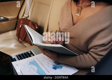Close-up of unrecognizable businesswoman in necklace sitting in car backseat and making notes in organizer while analyzing chart Stock Photo