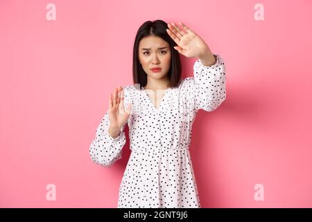 Timid and scared asian woman defending herself, raising arms in protection, victim being attacked, standing over pink background Stock Photo