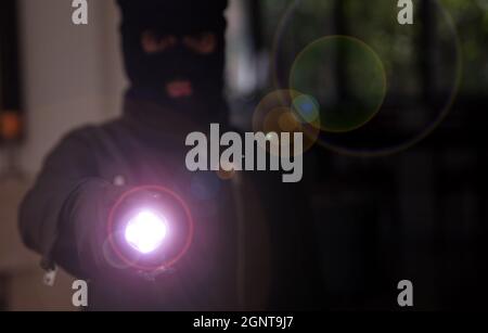 Housebreaking, burglary concept. Masked burglar in black balaclava entering a house holding a flashlight, dark night background. Stock Photo