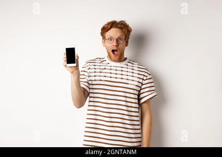 Technology and e-commerce concept. Surprised and shocked redhead guy checking out online promotion, showing blank smartphone screen and drop jaw Stock Photo