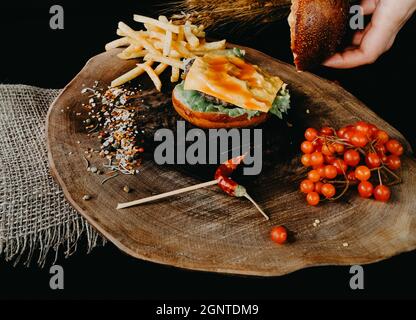 Sauce in hamburger on wooden plate with fries, small grains of spice and red currant on black background Stock Photo