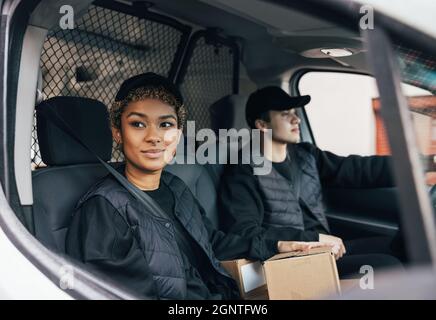 Two delivery coworkers in uniform sitting together in a van. Woman courier looking out from the window car while the man driving. Stock Photo