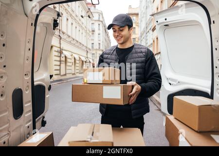 Smiling man courier in uniform standing at car trunk holding boxes preparing for delivery to a customer Stock Photo