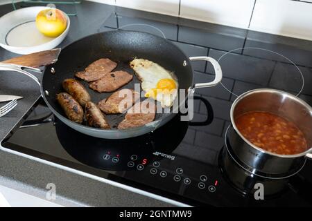 Traditional english breakfast in a kitchen with a fried egg, bacon rashers and sausages cooking in a frying pan, & baked beans in a saucepan. England Stock Photo