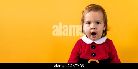 Banner Portraite Emotion Cute Cheerful Chubby Baby Girl in Santa Suit Yawns As If Singing At Orange Background. Child Christmas Scene Celebrating Stock Photo