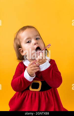 Portraite Cute Cheerful Chubby Baby Girl in Santa Suite Holding Eating Caramel Candy At Yellow Background. Child Play Christmas Scene Celebrating Stock Photo