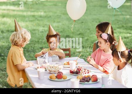 Portrait of cute curly boy opening gifts during outdoor Birthday party in Summer, copy space Stock Photo