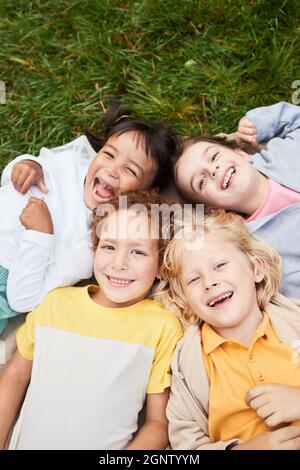 Vertical top view portrait of diverse group of kids lying on grass in park and looking at camera smiling Stock Photo