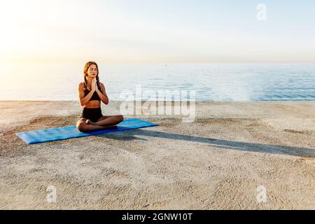 Young woman doing yoga by the sea. Sukhasana position Stock Photo