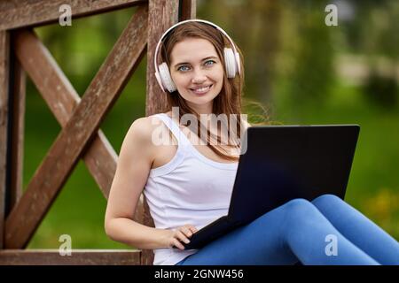 Smiling woman in headphones makes remote work with laptop outdoors. Stock Photo