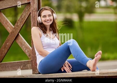 Smiling white woman in headphones sits in park in summer. Stock Photo