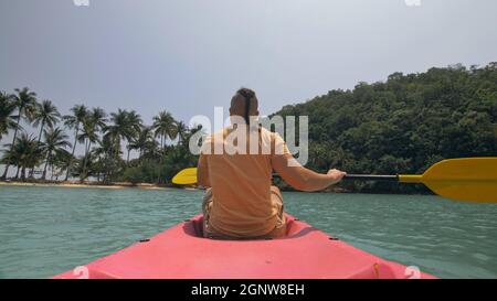Man with sunglasses and hat rows pink plastic canoe along sea against green hilly islands with wild jungles. Traveling to tropical countries. Stock Photo