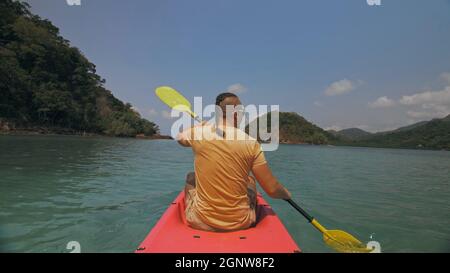 Man with sunglasses and hat rows pink plastic canoe along sea against green hilly islands with wild jungles. Traveling to tropical countries. Stock Photo