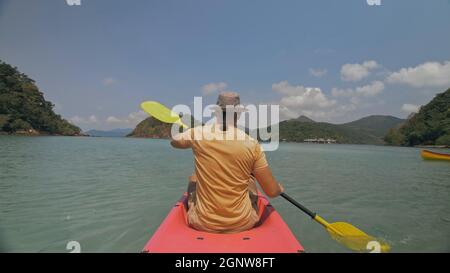 Man with sunglasses and hat rows pink plastic canoe along sea against green hilly islands with wild jungles. Traveling to tropical countries. Stock Photo