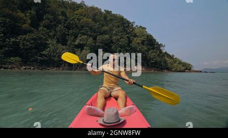 Man with sunglasses and hat rows pink plastic canoe along sea against green hilly islands with wild jungles. Traveling to tropical countries. Stock Photo