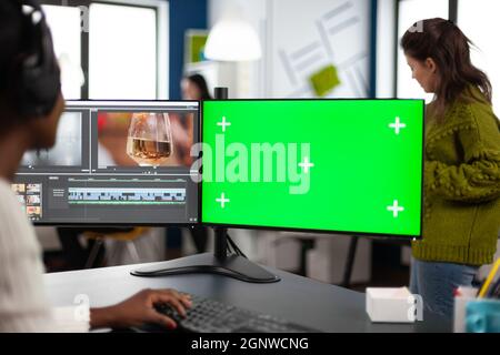 African designer video editor with headset editing footage using pc with greenscreen, chroma key isolated display sitting in agenc company. Videographer in digital multimedia company processing movie Stock Photo