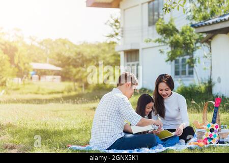 Happy Asian young family father, mother and child little girl having fun and enjoying outdoor on picnic blanket reading book in park at sunny time, su Stock Photo