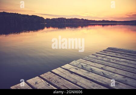 Sunset over still lake, wooden pier in foreground, color toning applied. Stock Photo
